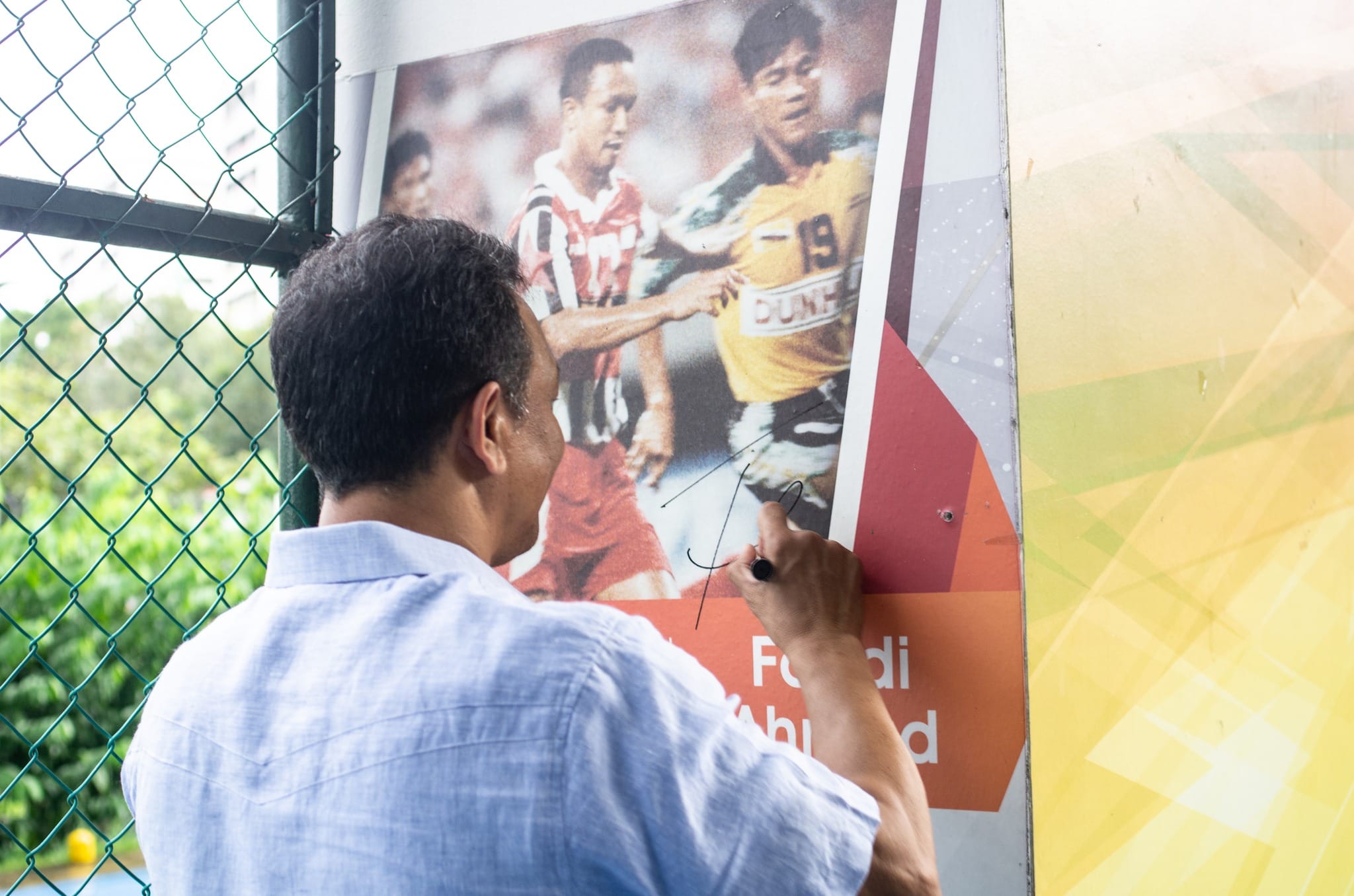 Man in blue shirt signing a poster showing two soccer players, one in red and one in yellow jersey number 19.